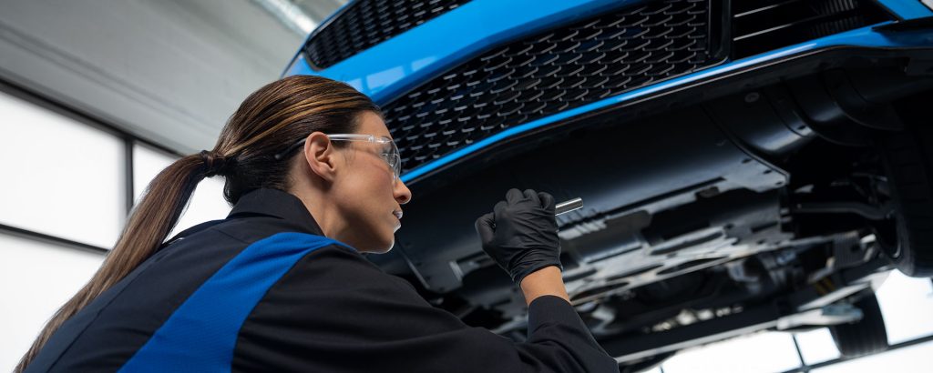Technician inspecting underside of a blue vehicle with a flashlight in a service bay.