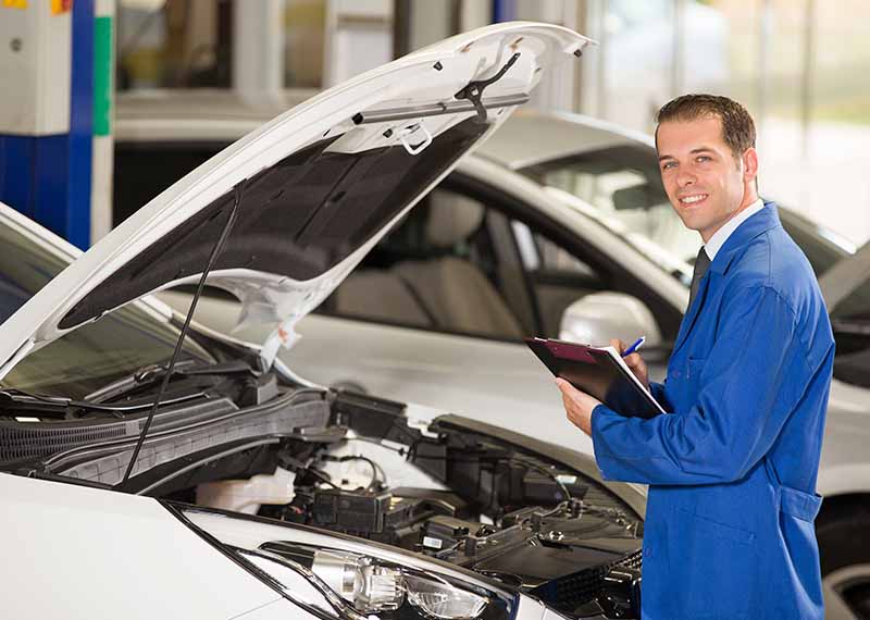 Auto mechanic in blue coveralls inspecting car engine with a clipboard in a service garage, smiling at the camera.